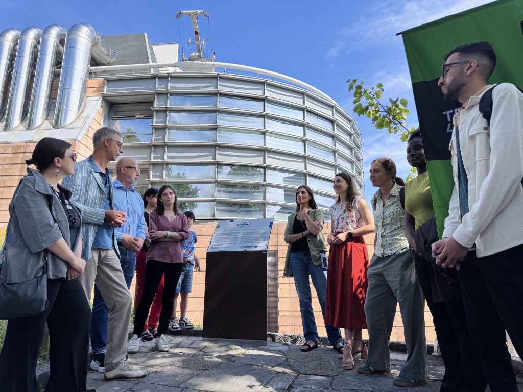 A group of people stands in a semicircle outdoors, listening to a man speak near a modern building with metal and glass features. The group appears engaged, with a plaque and a green banner visible. The sky is clear and sunny.