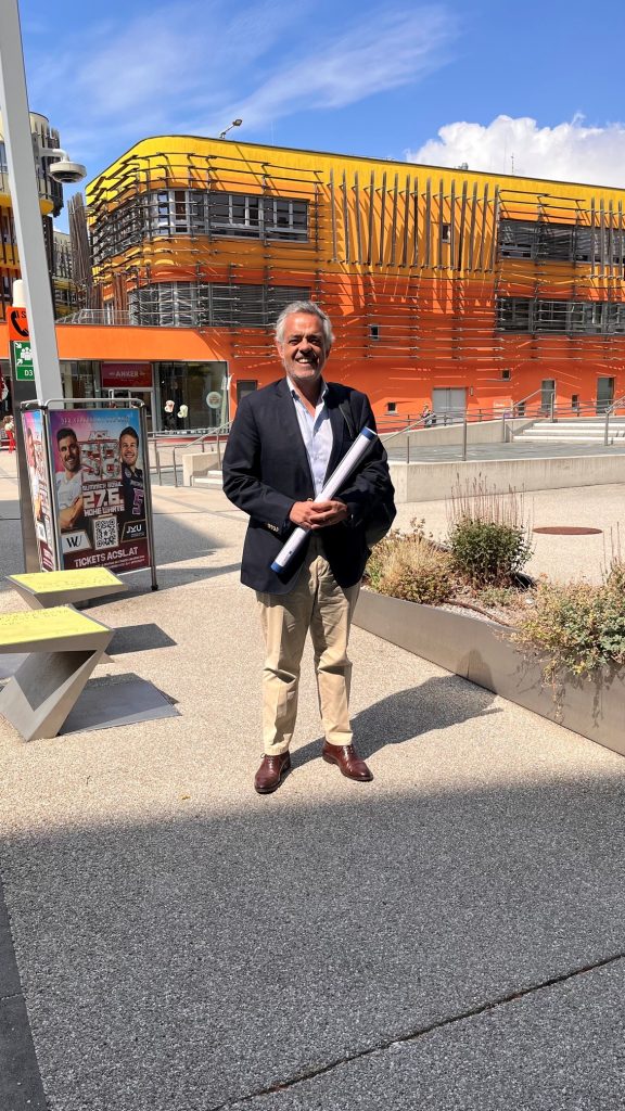 A man in a navy blazer and beige pants stands outdoors holding rolled papers, smiling. Behind him is a modern orange and yellow building and a poster on a bus stop. The sky is blue with some clouds.