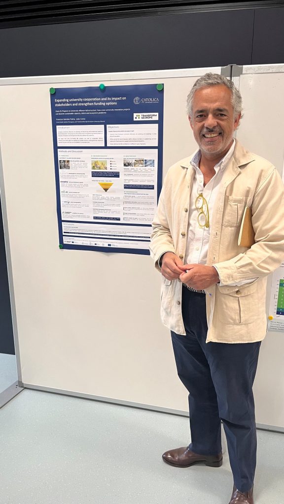 A man stands smiling next to a research poster displayed on a whiteboard. He is wearing a beige jacket, holding glasses, and has a pen in his pocket. The poster contains academic content with text, images, and charts.