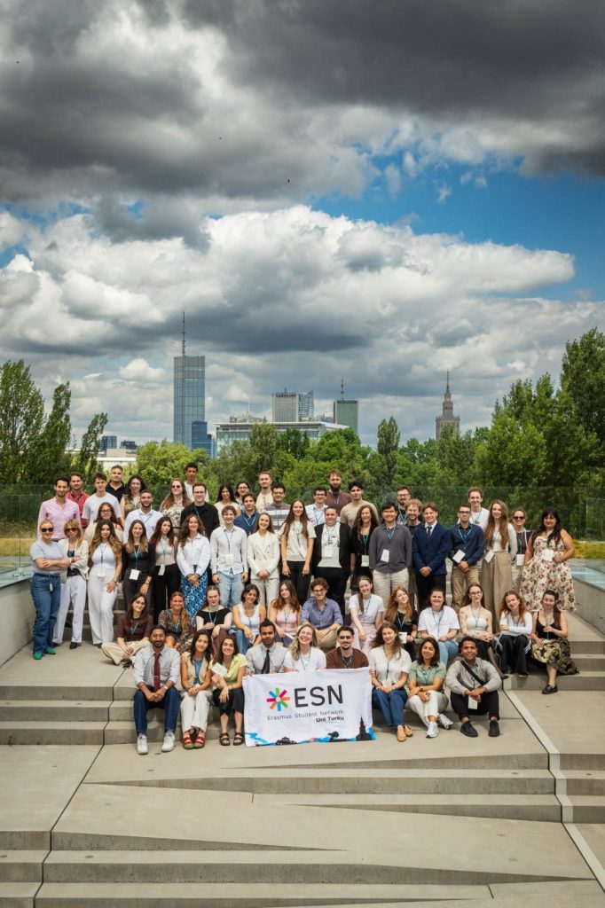 A large group of people pose on outdoor steps, holding a banner that reads ESN. Trees and city buildings are visible in the background under a partly cloudy sky.