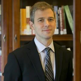 A man in a suit and tie stands in front of a bookshelf filled with books, looking at the camera with a slight smile.