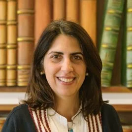 A woman with shoulder-length dark hair smiles, wearing a white shirt with a red and black patterned sweater. She stands in front of a bookshelf filled with large, colorful books.