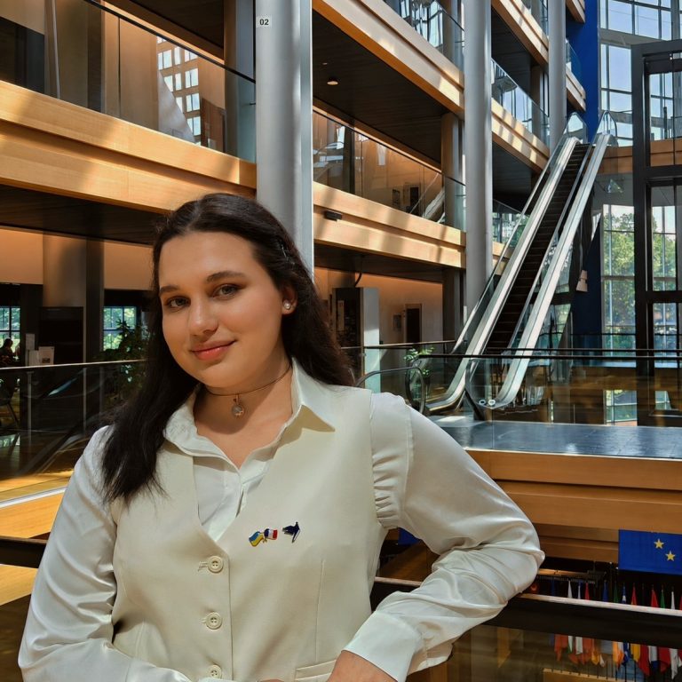 A woman with long dark hair, wearing a white blouse and vest adorned with small flag pins, stands indoors in a modern building—with glass railings, wood accents, and escalators in the background—embodying the spirit to Transform for Europe.