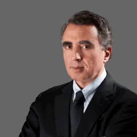 A man in a dark suit and tie poses against a plain gray background, looking at the camera with a neutral expression and his arms crossed.