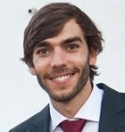 A young man with brown hair and a beard, wearing a dark suit, white shirt, and red tie, smiling at the camera against a light background.