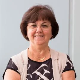 A woman with short brown hair, glasses, and a patterned top smiles while standing with arms crossed in front of a plain light-colored background.