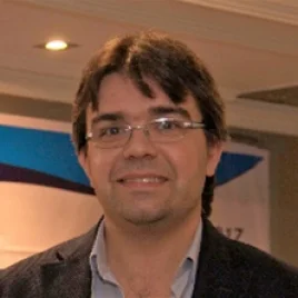 A man with glasses, dark hair, and a slight smile is wearing a blazer and shirt, standing indoors with a blurred colorful banner in the background.