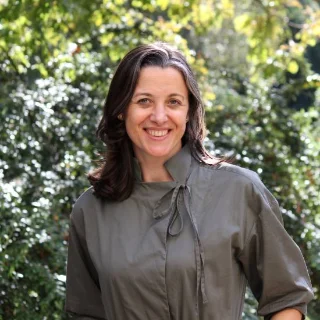 A woman with shoulder-length brown hair, wearing a gray collared shirt, stands outside and smiles at the camera. Green leafy trees fill the background.