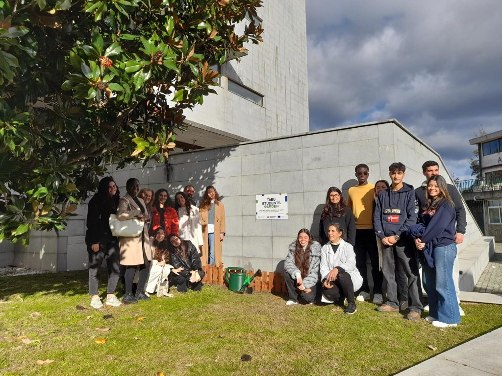 A group of young people pose together outside a modern building, standing and kneeling on grass beside a small wooden fence and a sign that reads The Green School Project.