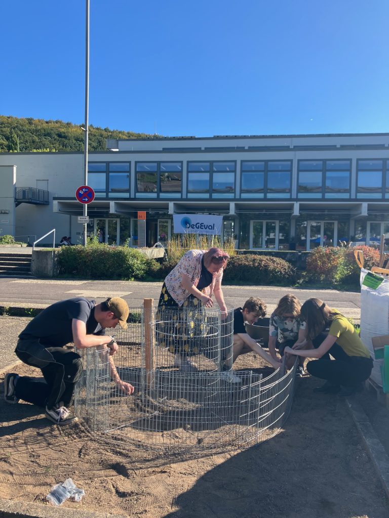 Five people are assembling a circular wire fence structure on a sandy area outside a modern building on a sunny day. Lush green hills and a blue sky are visible in the background.