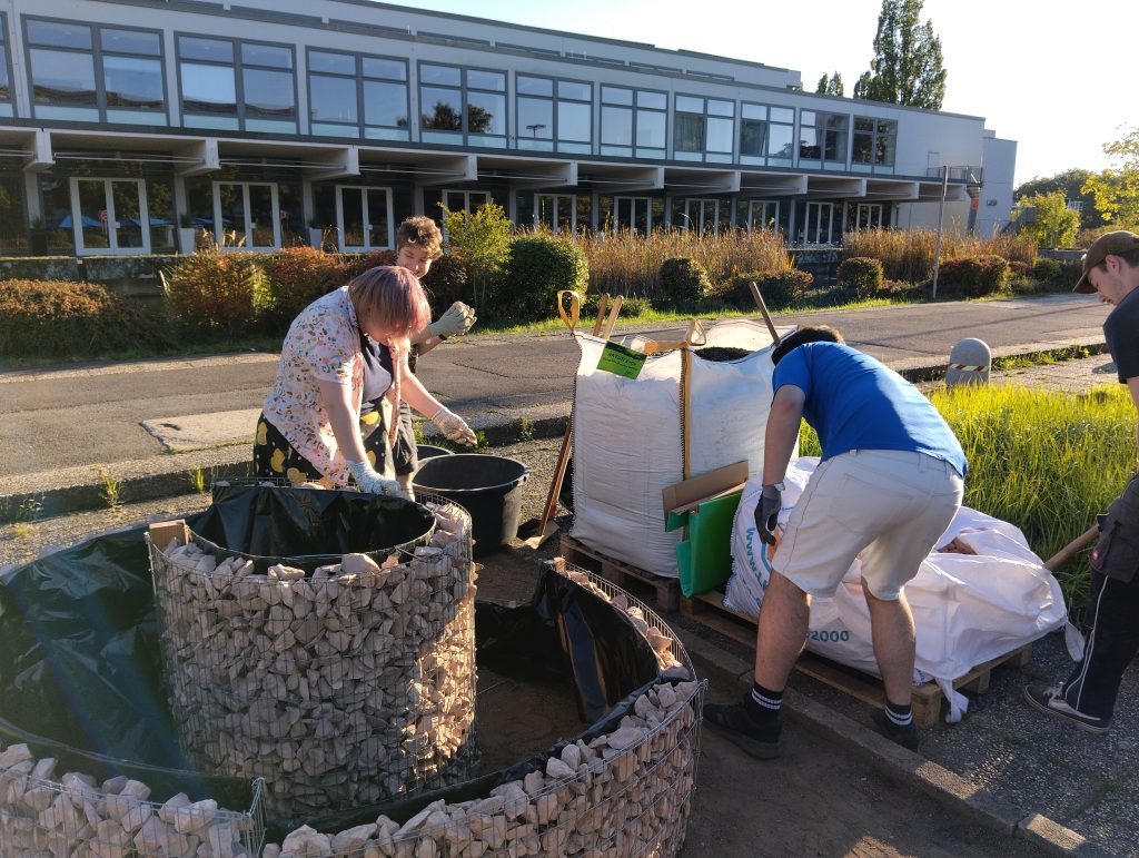 Four people are working outdoors, filling large wire mesh and plastic containers with rocks and soil near a modern building on a sunny day. Bags of materials and gardening tools are scattered around them.