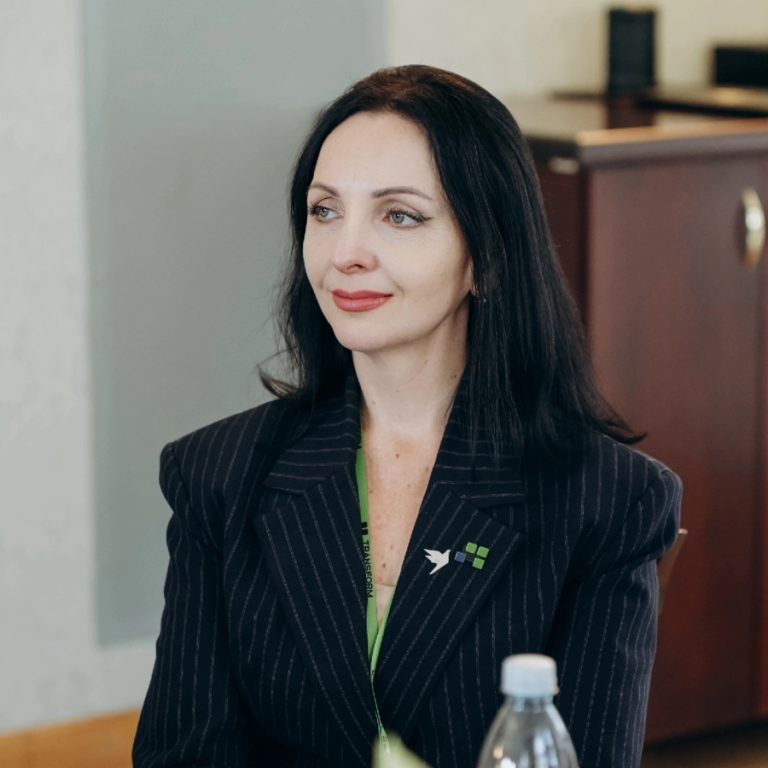A woman with long dark hair, wearing a pinstripe blazer and a green top, sits at a table with a water bottle in front of her. She is looking slightly to the side and smiling gently, embodying the spirit of Transform for Europe as she participates in this collaborative environment.
