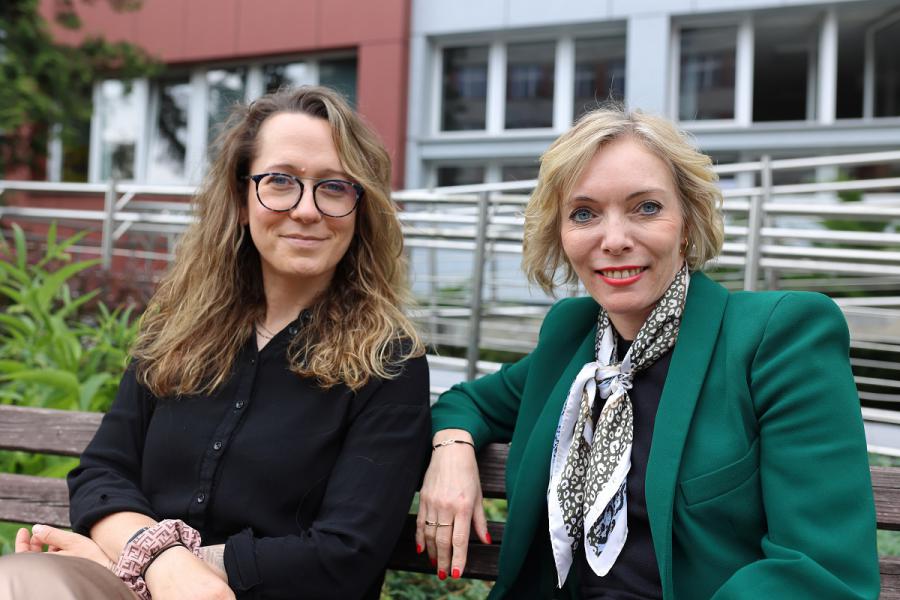 Two women sit on a bench outdoors, smiling at the camera. One wears glasses and a black shirt, the other has a green blazer and a patterned scarf. Modern buildings and greenery are in the background.