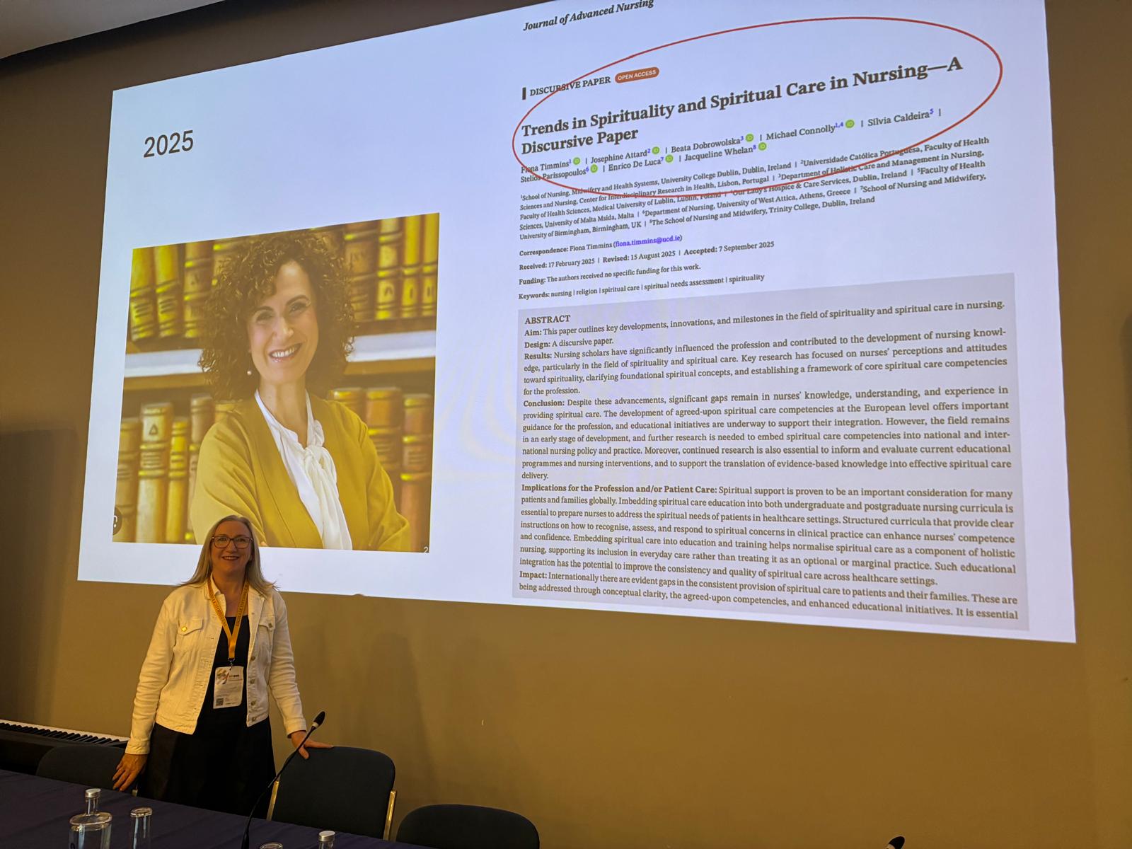 A woman stands in front of a large projected screen showing a journal article titled Trends in Spirituality and Spiritual Care in Nursing—A Discursive Paper and a photo of a smiling woman with curly hair.