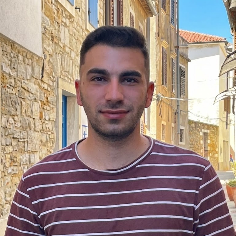 A man in a maroon and white striped shirt stands in a narrow street lined with stone buildings and colorful shutters, beneath a bright blue sky—capturing the vibrant atmosphere of cities that Transform for Europe.