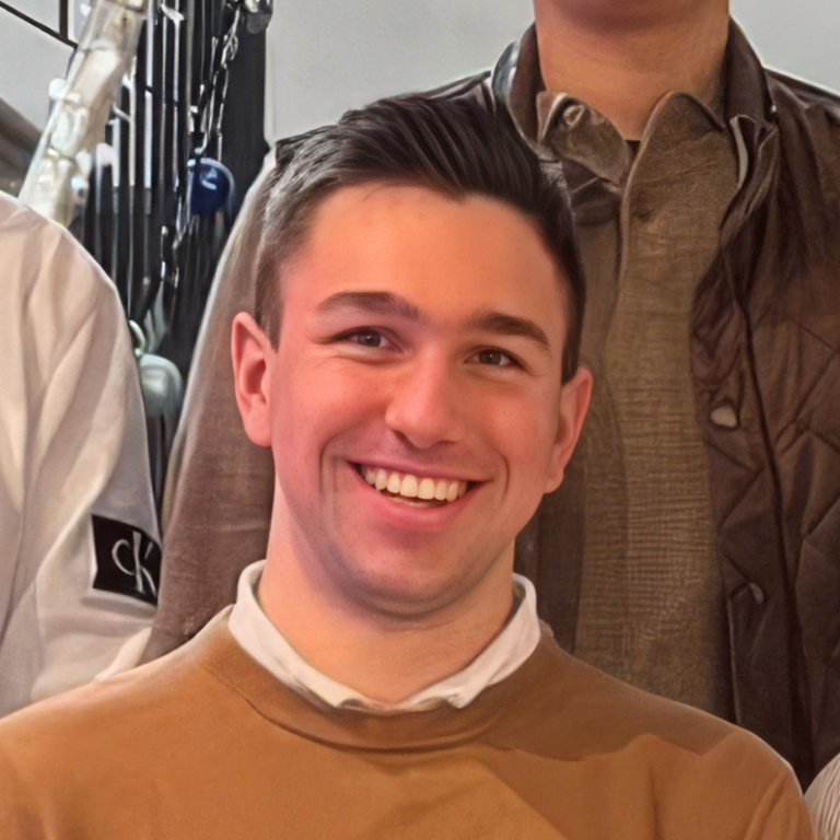 Young man with short dark hair smiles at the camera, wearing a tan sweater over a white collared shirt. Representing the spirit of Transform for Europe, two people in darker clothing stand partially visible beside and behind him. Indoor setting with blurred background.