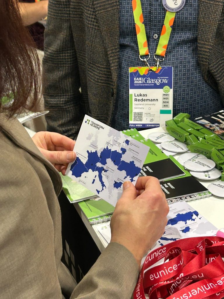 A person holds a map with blue regions at a conference booth. Another person wears a badge reading Lukas Redemann, EAIE Glasgow 2024. Lanyards, pins, and brochures are visible on the table.