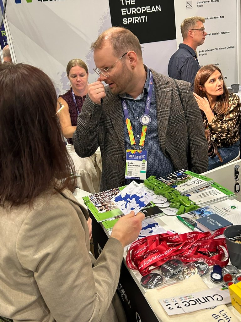 A man and woman talk at a busy booth with brochures, lanyards, and promotional items on the table. Other people are visible in the background. The setting appears to be a conference or expo.