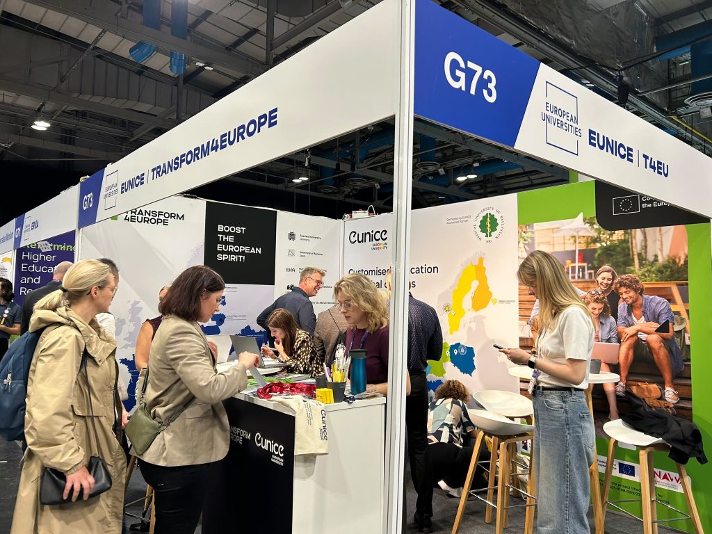 People interact at a busy exhibition booth labeled TRANSFORM4EUROPE and EUNICE with maps and group photos on display, under bright overhead lights in a large event hall.