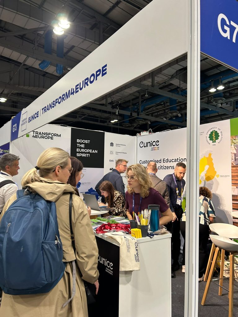 People gather at the EUNICE Transform4Europe booth at an expo. Attendees interact with staff, pick up informational materials, and explore displays under bright lighting in a modern exhibition hall.