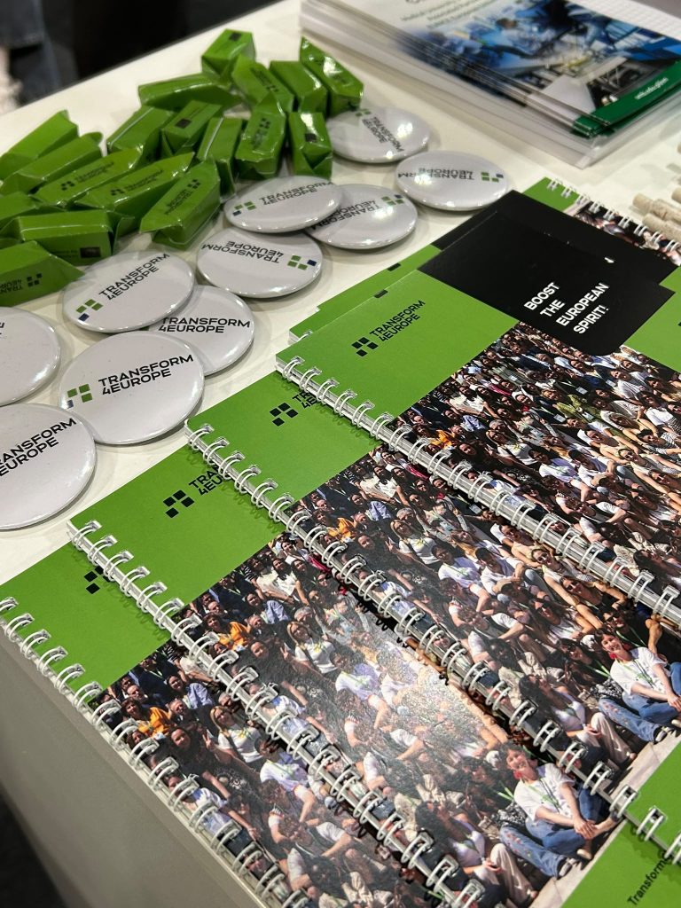 Green notebooks, white Transform Europe buttons, and green USB drives are arranged on a table with some pamphlets in the background. The notebooks have a group photo and event branding on the cover.