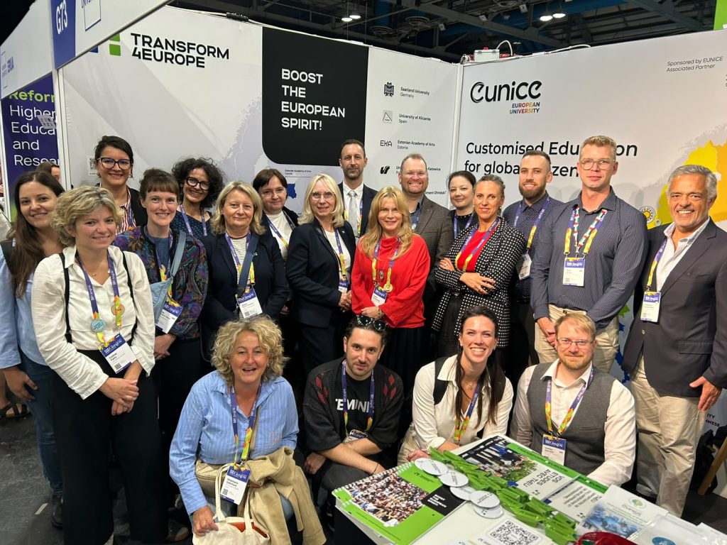 A group of people pose and smile at a trade show or conference booth displaying banners for Transform 4 Europe and CUNICO. Most wear badges and are standing or sitting at a table with brochures.
