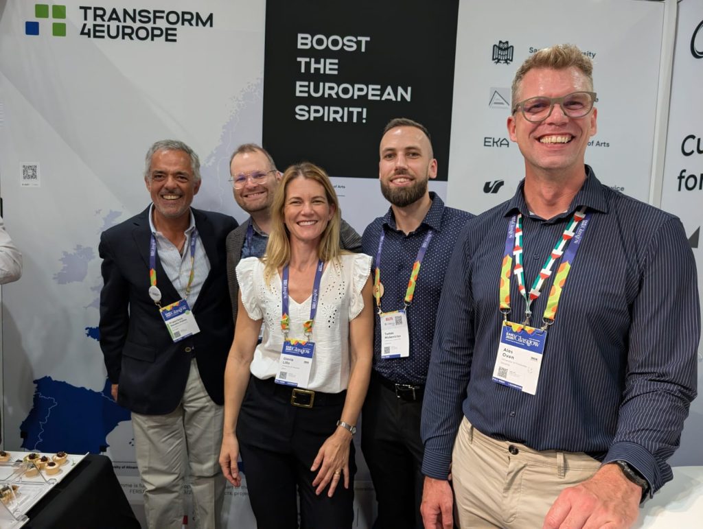 Five smiling people stand together at a booth with Boost the European Spirit! and Transform 4 Europe on the background, wearing conference badges and colorful lanyards. Some snacks are visible on the table.