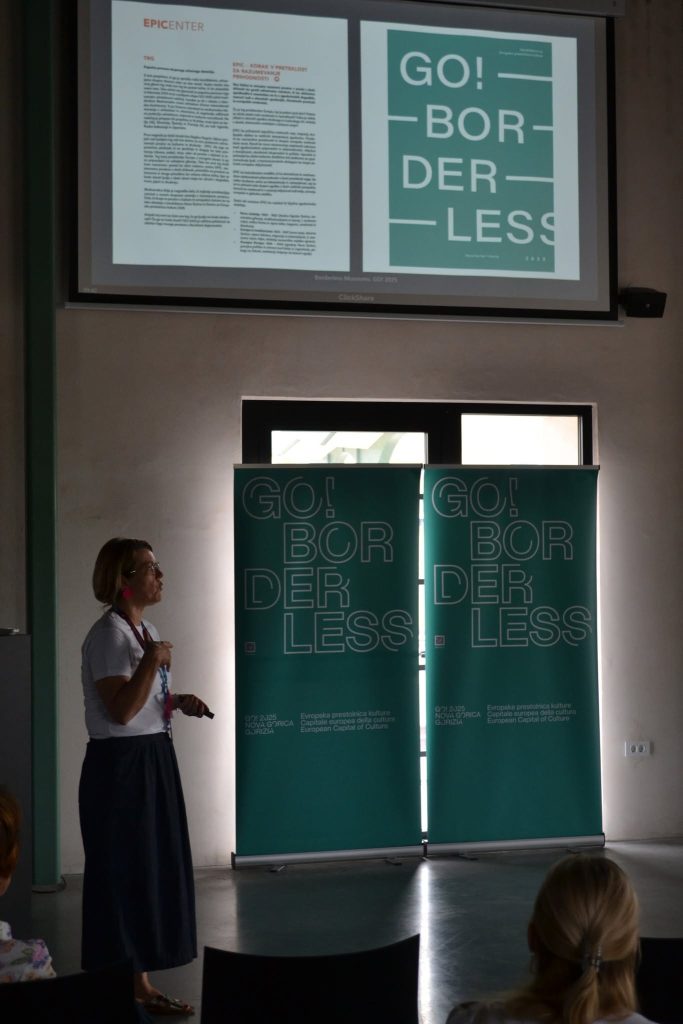 A woman presents in front of two green banners that read GO! BORDERLESS and a projection of similar materials in a dimly lit room with several attendees watching.
