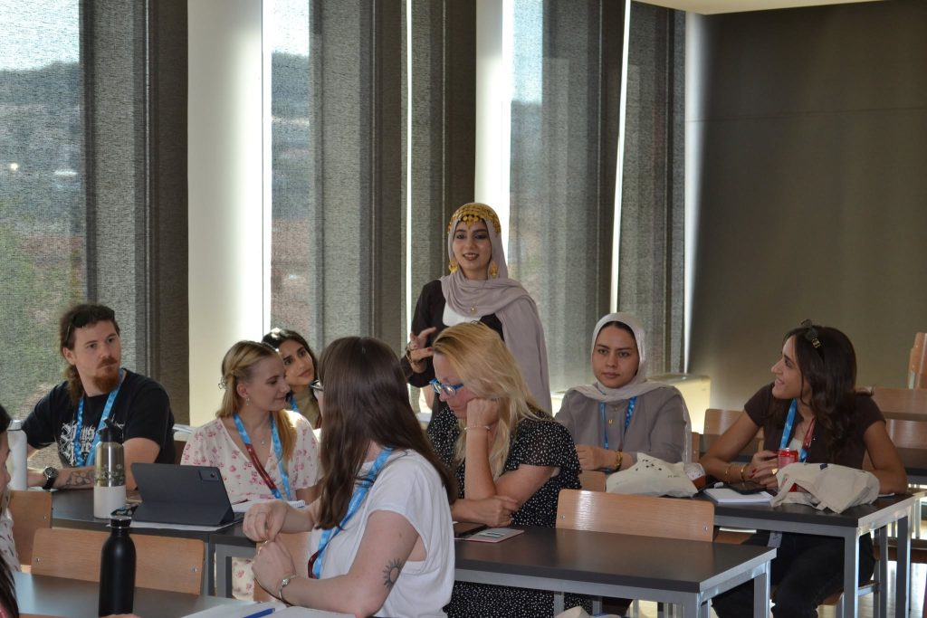 A group of people sit in a classroom, some talking to each other. One woman in a yellow headscarf stands and speaks, while others listen or look at their laptops. Sunlight filters through tall windows.
