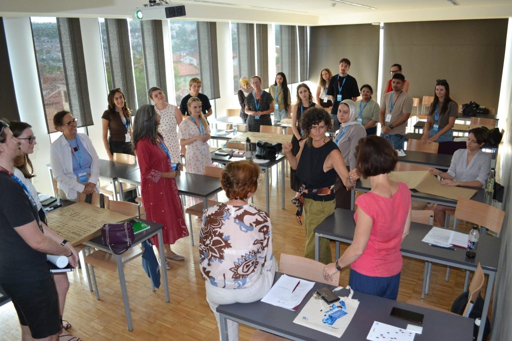 A group of people stands in a classroom, engaged in discussion around tables with papers and laptops. Some are seated while others stand, and natural light comes in through large windows.