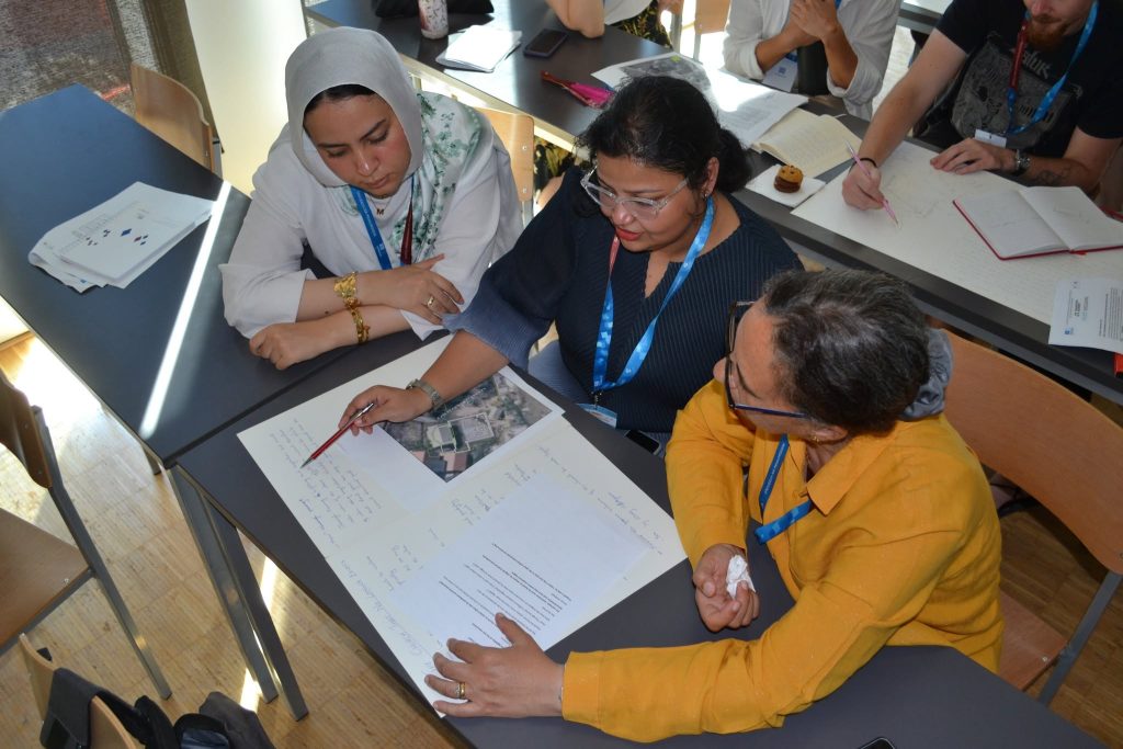 Three women sit at a table collaborating, reviewing notes and a map. They wear conference lanyards and are engaged in discussion, while others take notes nearby. Sunlight streams into the room.