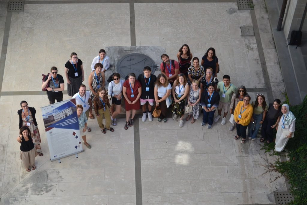 A group of people wearing name badges stands in a courtyard, looking up and smiling. Some hold papers, and theres a large poster board on the left side. The group is surrounded by stone tiles.
