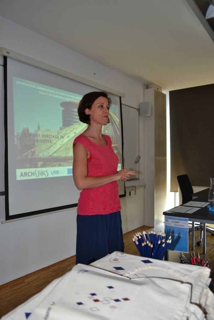 A woman in a pink top stands and speaks in a classroom. Behind her, a projector screen displays a presentation titled ARCHETHICS URB. A table with pencils and papers is in the foreground.