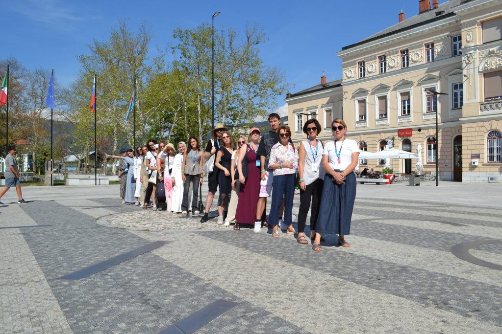 A group of people stands in a line on a sunny day in a town square, with buildings, flags, and trees in the background.
