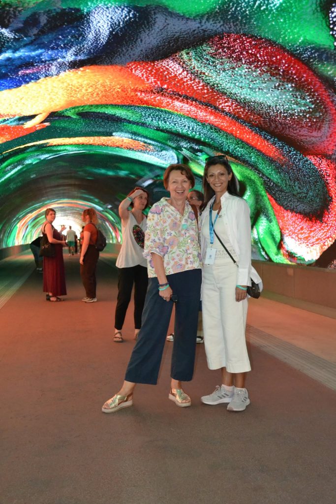Two women smile and pose together in a tunnel with vibrant, swirling, colorful patterns displayed on the walls and ceiling. Several other people are visible in the background of the tunnel.