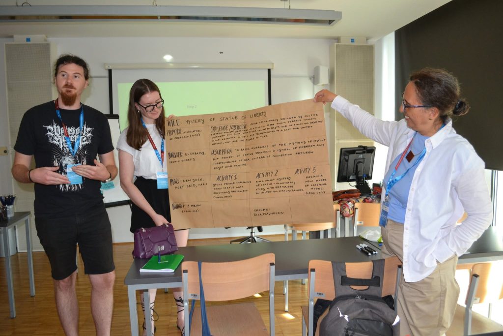 Three people stand in a classroom. Two people hold a large sheet of paper with handwritten notes, while another stands beside them speaking. Empty chairs and desks are visible, and a projector screen is in the background.