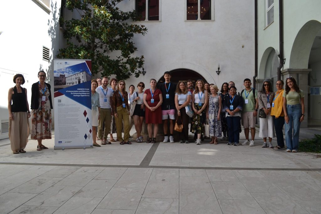 A group of people stands outdoors in a courtyard next to a large informational poster, smiling at the camera. The background features a white building with arched doorways and small trees.