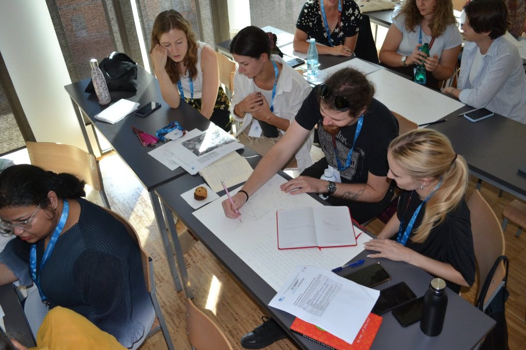 A group of people sit at tables, collaborating and writing notes on large sheets of paper during a workshop or classroom activity.