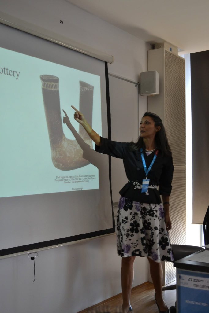 A woman stands and points at a projected slide showing ancient pottery while giving a presentation in a classroom.
