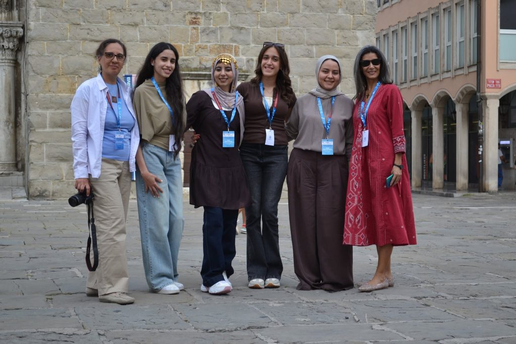 Six women wearing conference badges stand together outdoors on stone pavement, smiling for the camera. Some wear headscarves and casual clothes, one holds a camera, and the background includes stone buildings.