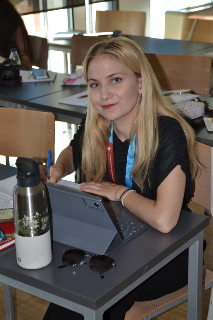 A young woman with long blonde hair sits at a desk, writing on paper next to a tablet, water bottle, and sunglasses in a classroom setting.