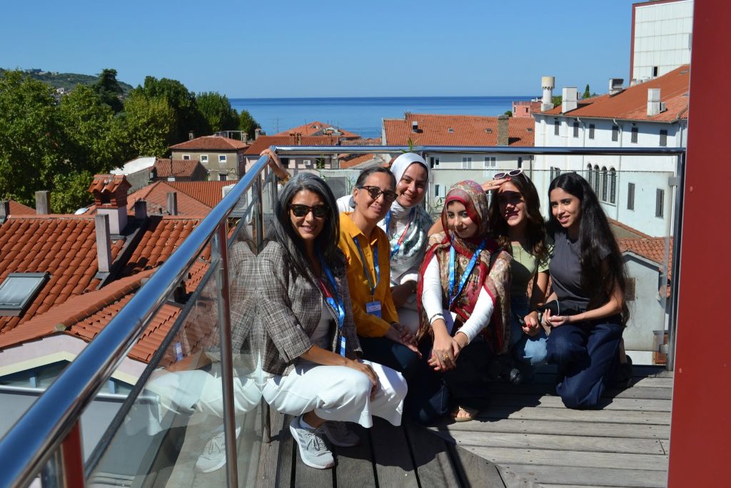 Six women smile and pose together on a rooftop terrace with red-tiled roofs, trees, and the sea in the background under a clear blue sky.