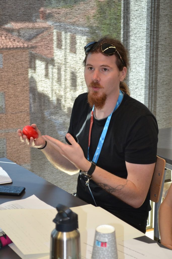 A person with sunglasses on their head holds a red object and gestures while sitting at a table with papers, a notebook, a metal bottle, and a paper cup. Sunlight streams through a window in the background.