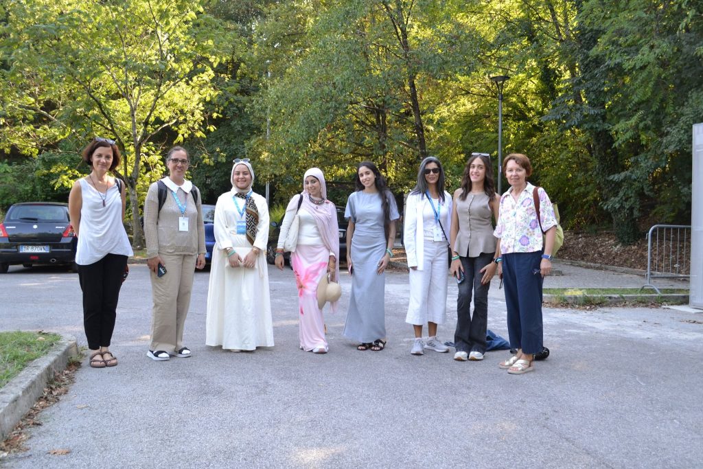 Eight women stand side by side outdoors on a paved path, surrounded by trees and greenery, smiling at the camera on a sunny day.