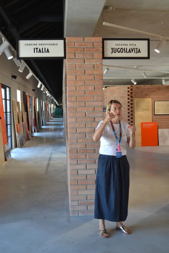 A woman stands indoors near a brick pillar beneath signs labeled Italia and Jugoslavia, gesturing with her hands in a museum or exhibition space with historical displays on the walls.