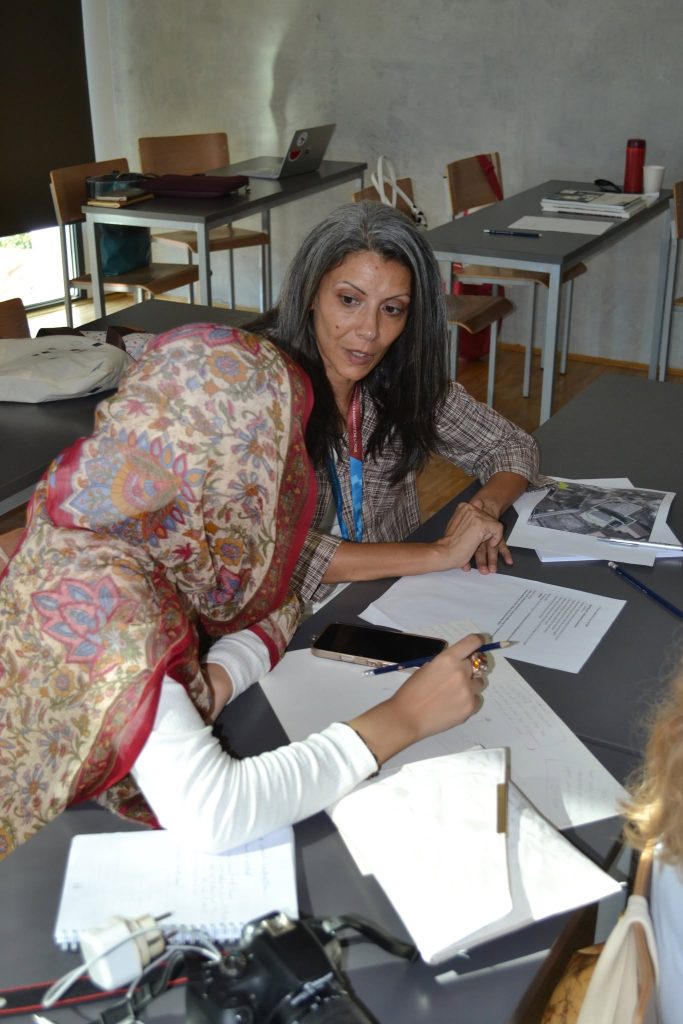 Two women sit at a table covered with papers, notebooks, a camera, and a phone. One wears a patterned scarf, and they appear to be discussing documents in a classroom setting.