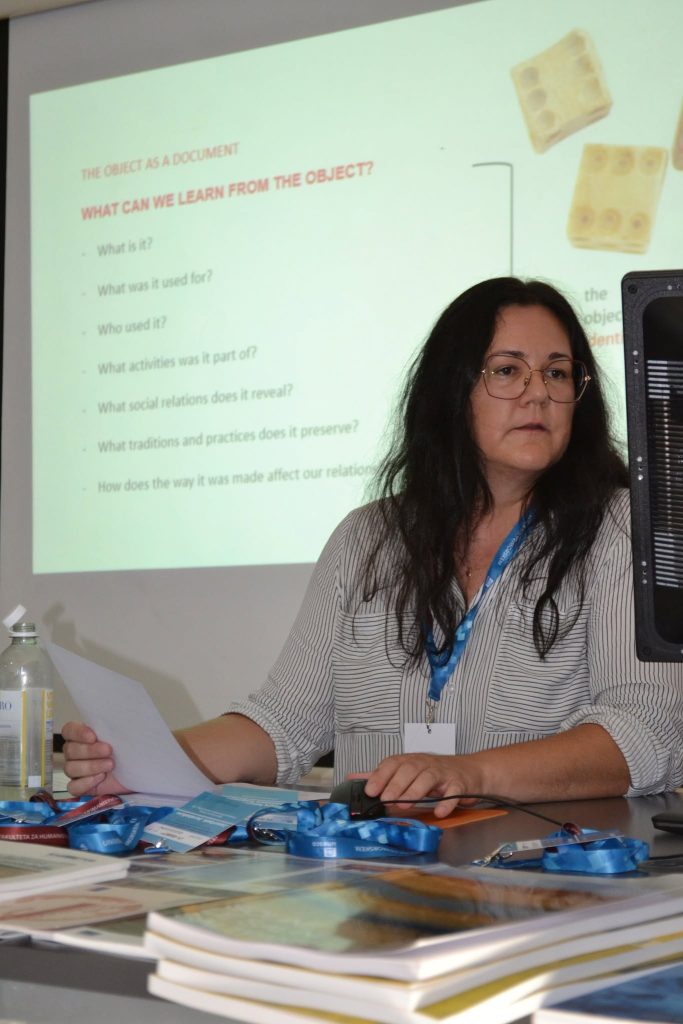A woman with long dark hair and glasses sits at a desk with papers and lanyards, presenting in front of a projected slide about learning from objects.