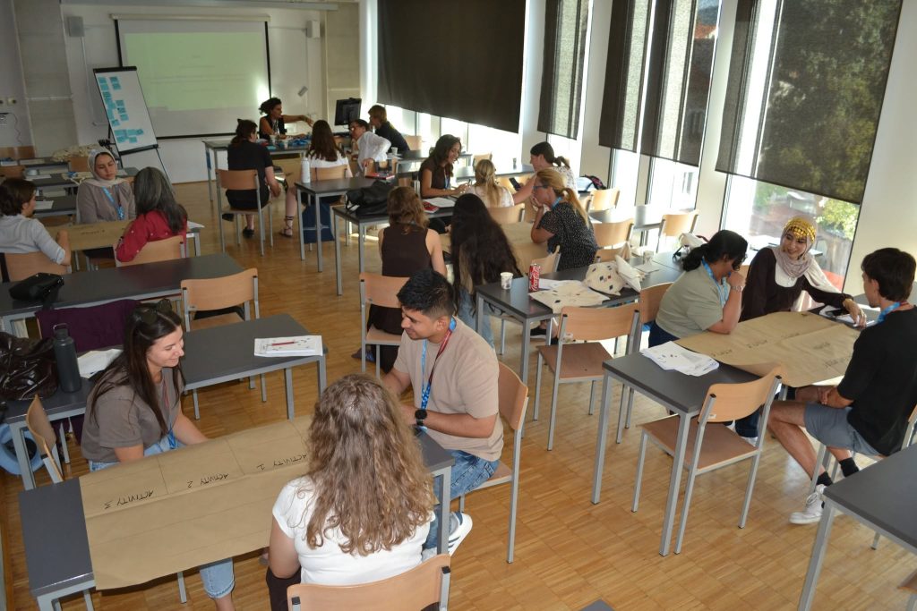 Students work in groups at tables in a bright classroom with large windows and a projection screen, actively discussing and writing on large sheets of paper.