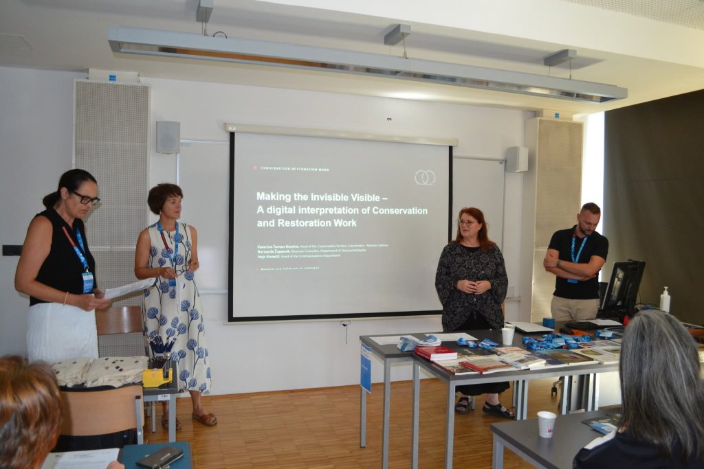 Four people stand in front of a projected presentation titled Making the Invisible Visible in a modern classroom, with desks, books, and an audience member visible in the foreground.