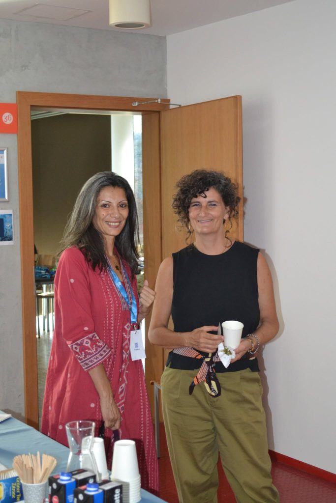 Two women stand by a door, one in a red patterned dress and badge, the other in a black top and olive pants holding a cup, both smiling. Table with drinks and cups is in the foreground.